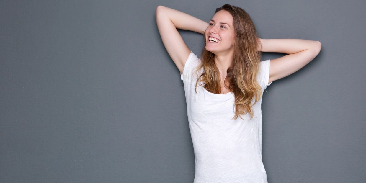 Happy woman in a white shirt smiling and relaxing against a gray wall.