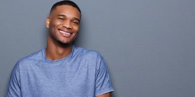Young man smiling and standing against a gray wall with hands in pockets.
