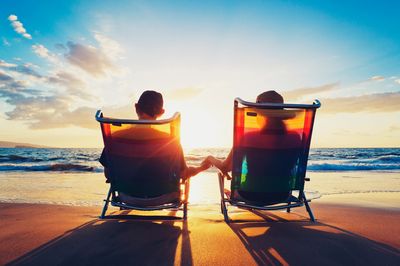 A couple sit on deckchairs hand in hand on the beach at sunset.