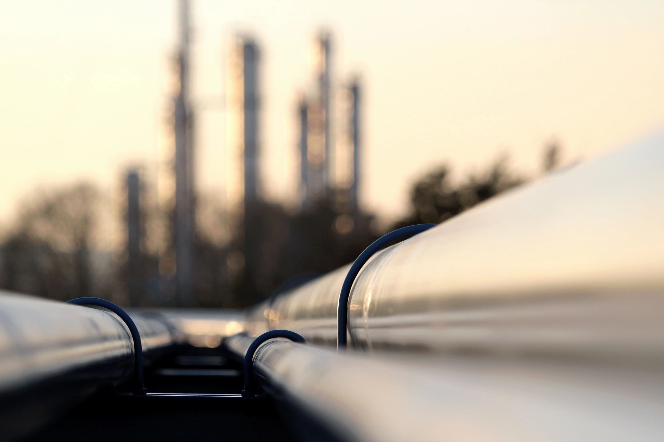 Close-up of industrial pipelines with a blurred refinery background at sunset.
