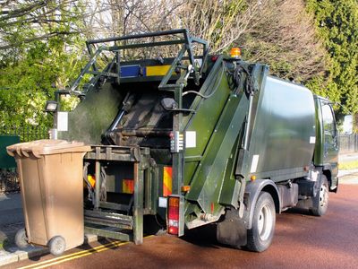 A green garbage truck emptying a brown trash bin on a suburban street.
