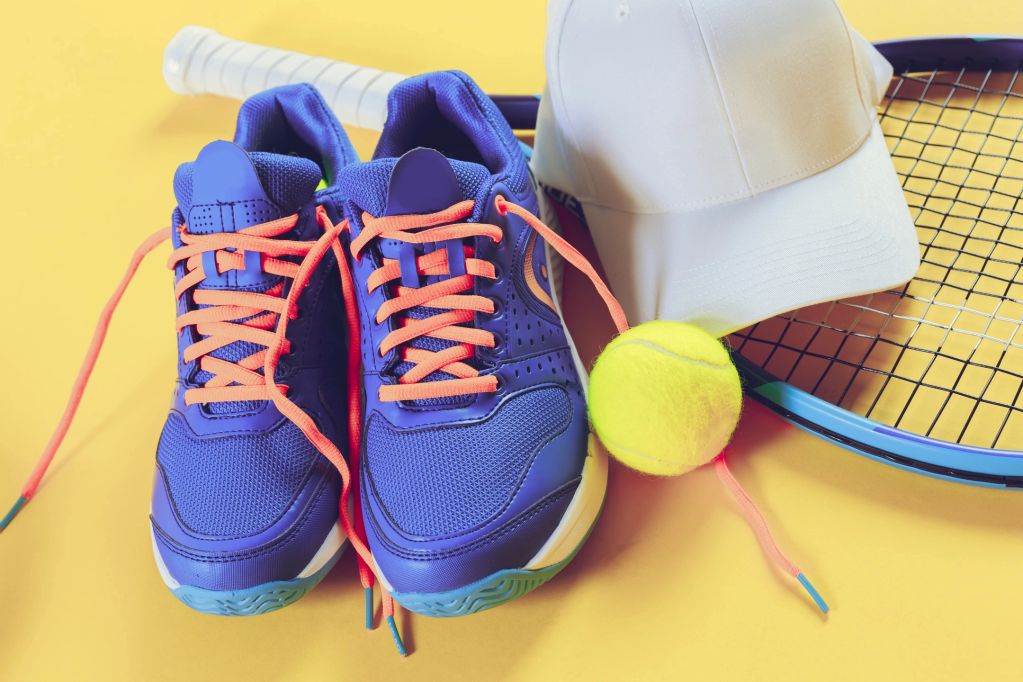 Blue tennis shoes, white cap, tennis ball, and racket on a yellow background.