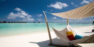 A hammock on a white sandy beach with colorful pillows under a shaded canopy.