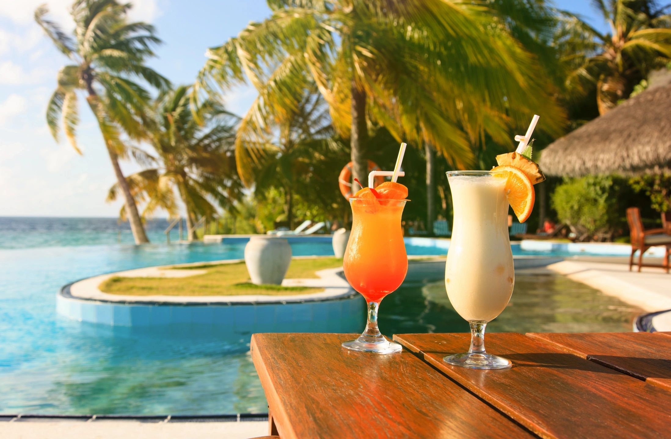 Two tropical cocktails by a pool with palm trees and ocean in background.
