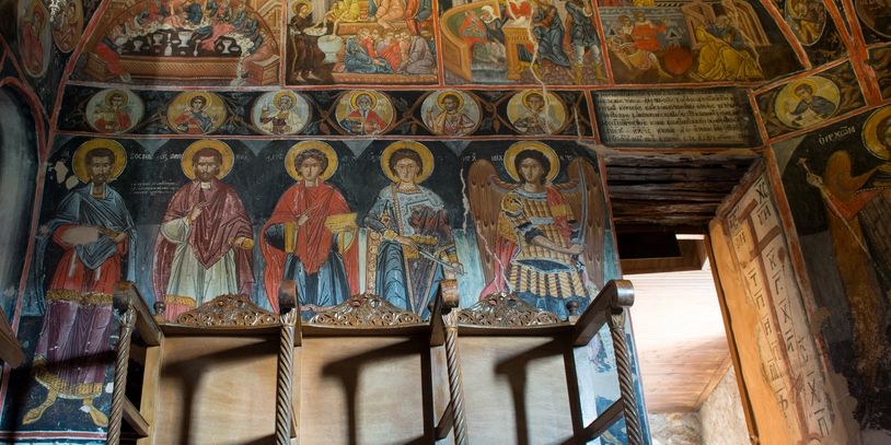 Interior wall frescoes of saints in a historic church with wooden chairs.