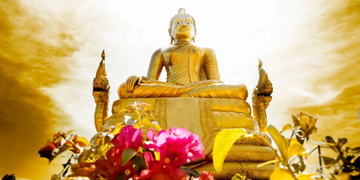 Golden Buddha statue with vibrant flowers in foreground under a dramatic sky.