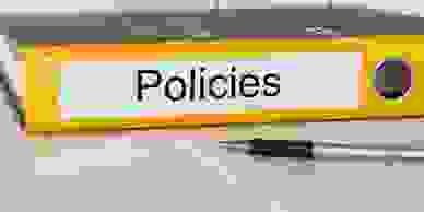 Yellow ringbinder folder resting on its side with the word "Policies" on its spine in front of a pen