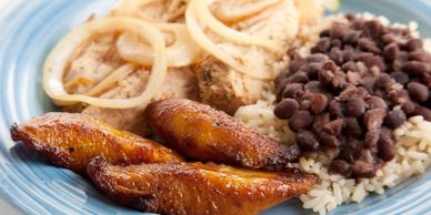 A plate with cooked plantains, rice with black beans, and seasoned chicken with onions.