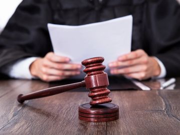 upper torso of man in black robe holding documents in his hands with gavel and table in front of him