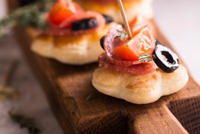 Mini flatbread appetizers with tomato, olive, and salami on a wooden board.