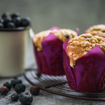 Blueberry muffins with crumb topping in purple wrappers, fresh blueberries nearby.