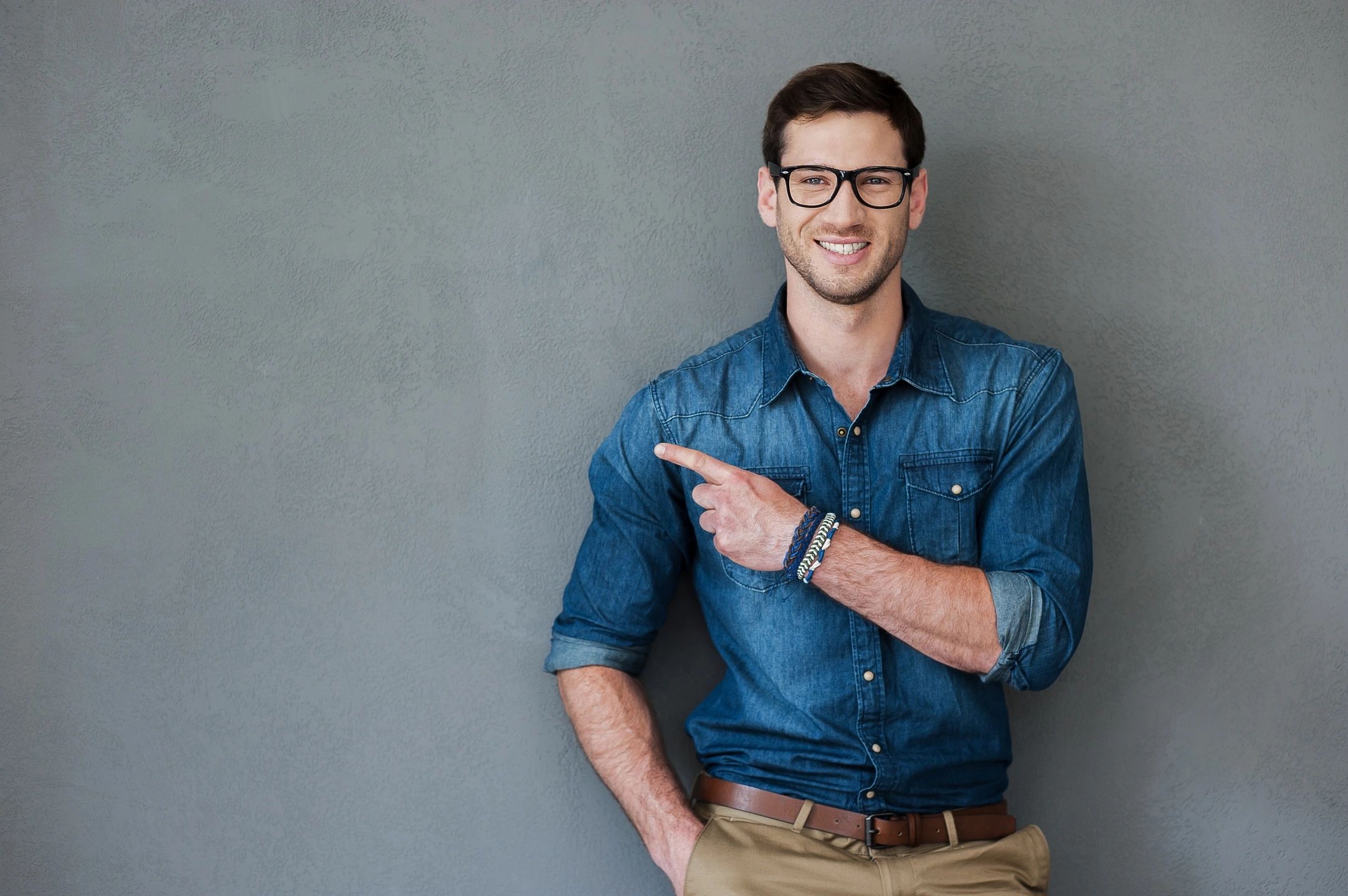 Smiling man in glasses pointing to the side against a gray wall.