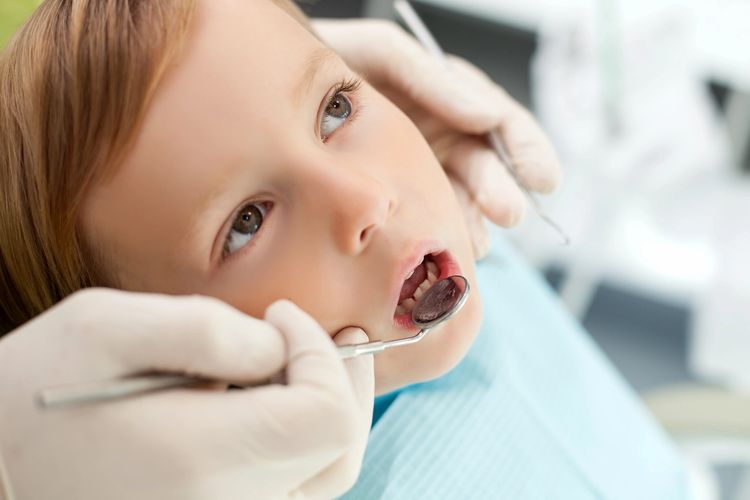 A child with mouth open getting an exam with a mouth mirror by a pair of gloved hands