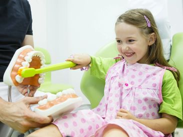 A smiling girl learns to brush teeth using a dental model and oversized toothbrush.