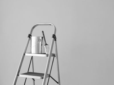 A paint can and brush on a metal step ladder against a plain wall.