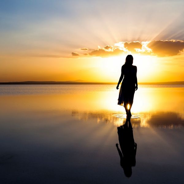 Silhouette of a woman walking on water at sunset with vibrant sky reflection.