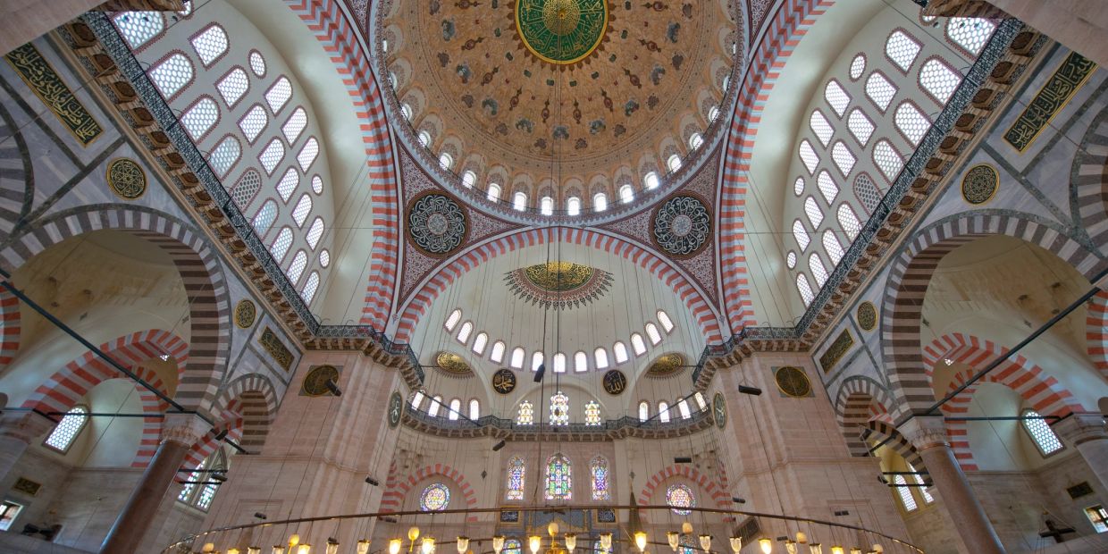 Intricate interior dome of a mosque with Islamic calligraphy and stained glass windows.