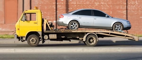 A silver sedan on a yellow tow truck against a brick wall backdrop.