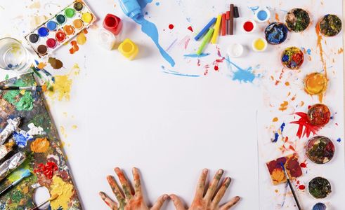 Hands covered in paint surrounded by art supplies on a white table.