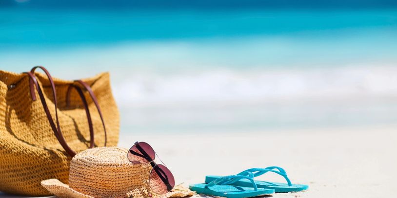 Beach essentials including a straw hat, sunglasses, bag, and blue flip-flops on sand.