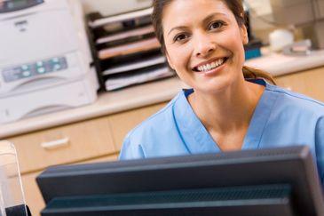 Smiling healthcare professional in blue scrubs at a computer desk.