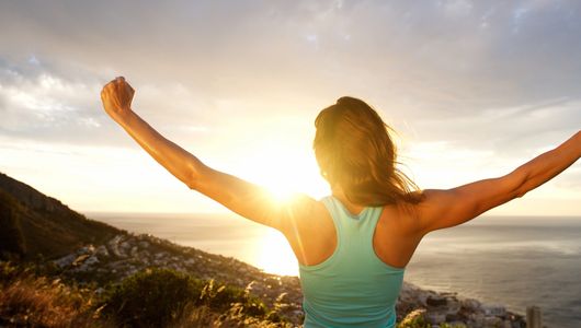 Woman looking over the ocean with arms raised and fist clenched like she just accomplished a victory