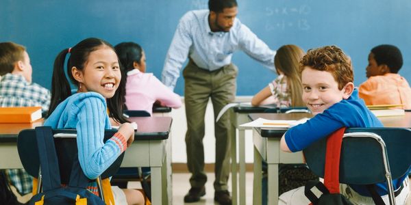 Classroom with teacher and students. Two students in focus turned around smiling at camera.