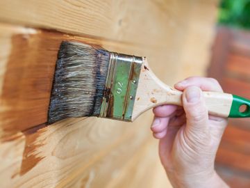 Hand applying wood stain with brush on wooden surface.