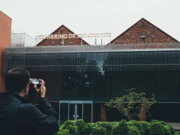 Person photographing a building with 'Gathering of Strangers' sign.