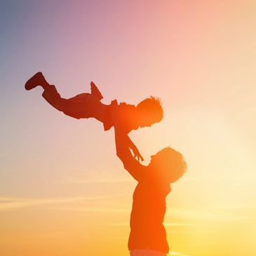 silhouette of a parent holding a child up in the sky as they smile at each other