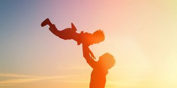 Silhouetted adult lifting child joyfully against a sunset sky.