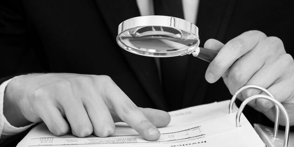 Security assessor with magnifying glass reviewing a binder.