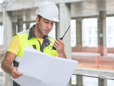 Construction worker in safety gear using a walkie-talkie and blueprint.