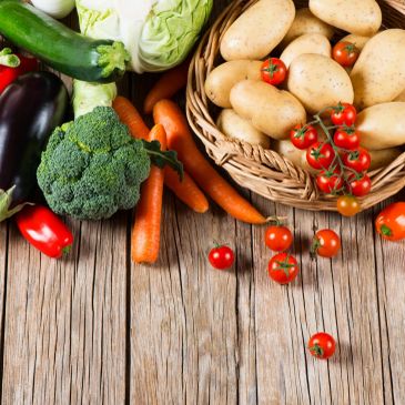 A basket of potatoes and various fresh vegetables on a wooden surface.