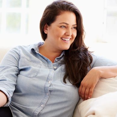 woman in long dark hair sitting on a white couch in front of a window, looking to the side smiling