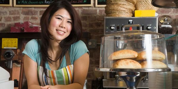 Smiling barista behind counter with bread rolls under a glass dome.