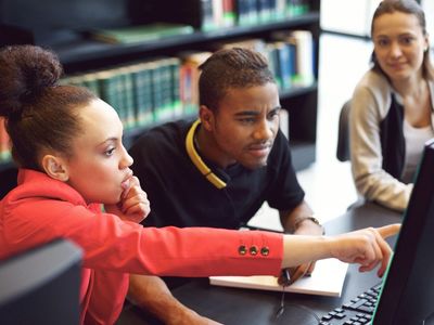 Three young adults collaborating and pointing at a computer screen in a library.