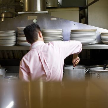 Chef stirring a pot in a kitchen with stacked plates above.