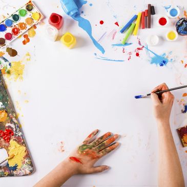 Artist hands messy with paint, shown hovering above a blank canvas with paints all around it