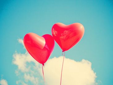 two heart shaped balloons nuzzling against each other, floating above fluffy white clouds 