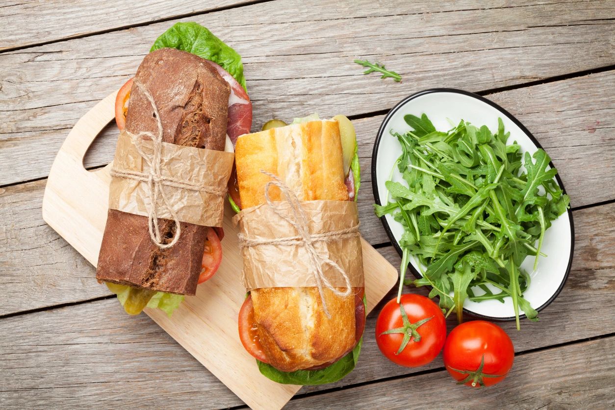 Two wrapped sandwiches on a wooden board with fresh arugula and tomatoes.
