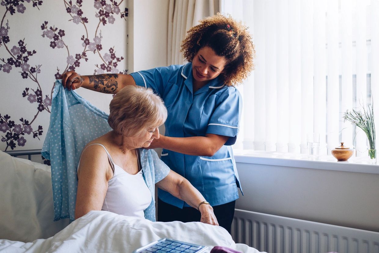 A caregiver helps an elderly woman put on a shirt in a cozy bedroom.