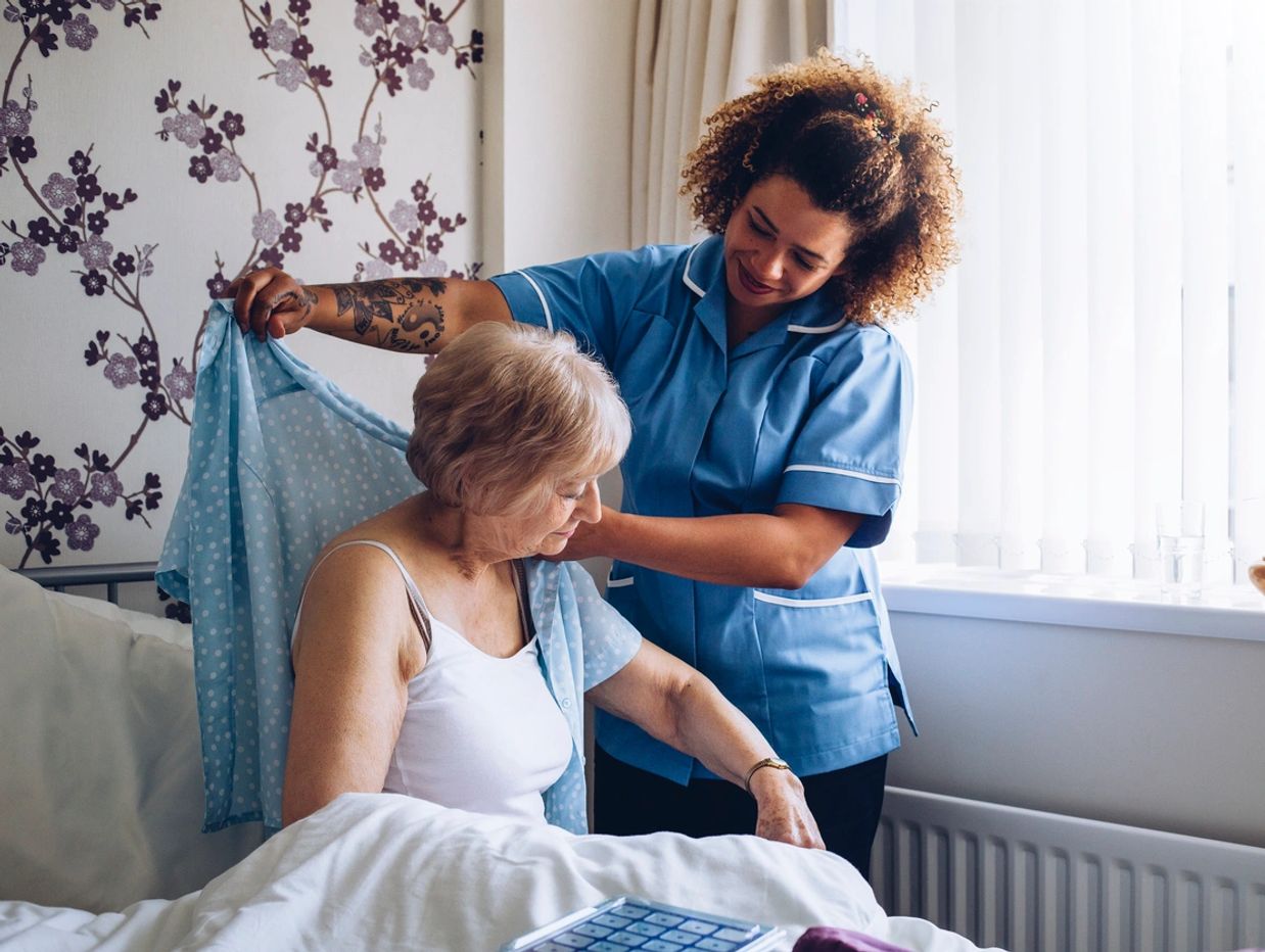 A nurse helps an elderly woman put on a blue shirt by her bedside.