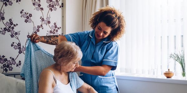 Caregiver helps elderly woman put on a light blue gown in a cozy room.