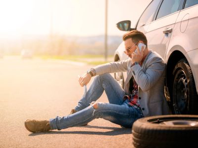 Man sitting by car with a flat tire, making a phone call for help.