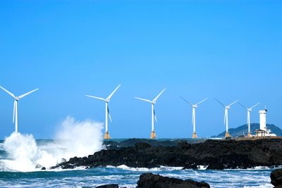 Wind turbines line a rocky coastline with waves crashing nearby under a clear blue sky.