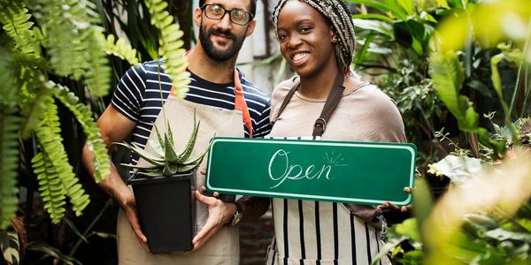 Two florists holding an 'Open' sign and a plant, smiling in their garden shop.