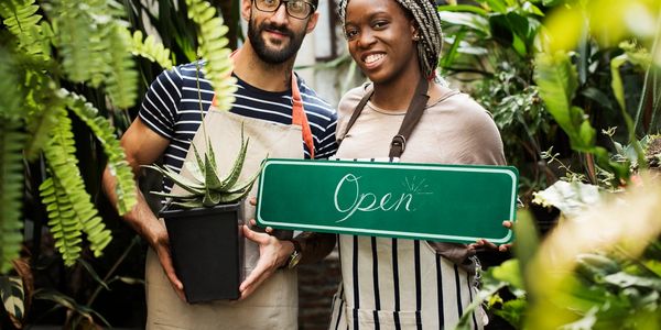 Two smiling florists holding an open sign and a potted plant in a lush garden shop.
