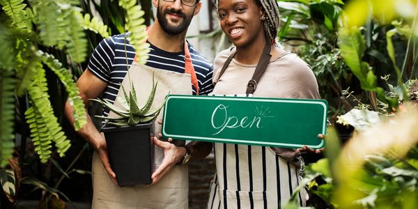 Two smiling florists holding an "Open" sign in a lush plant shop.