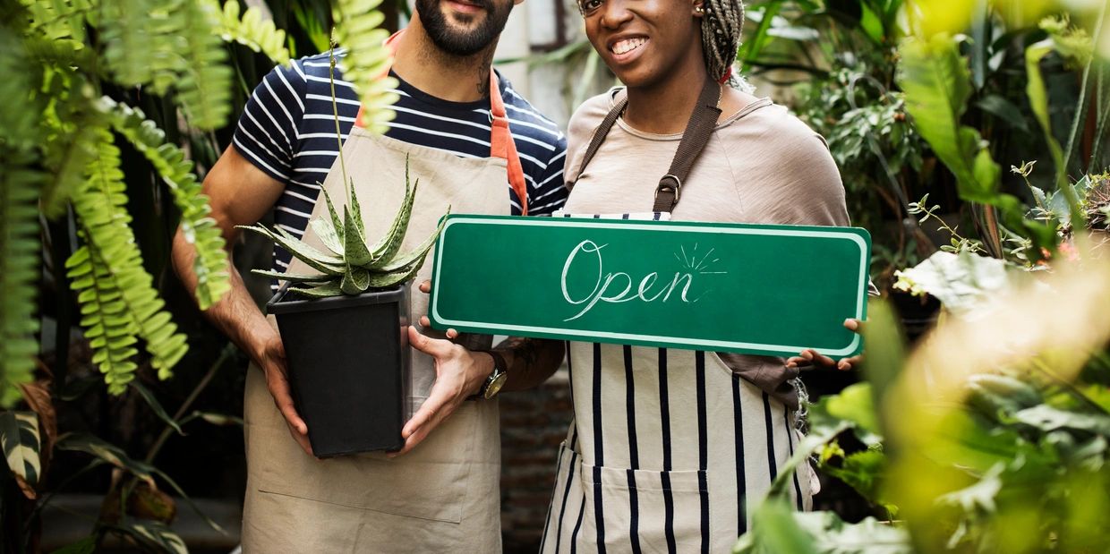 Two people with a plant and an open sign in a garden.
